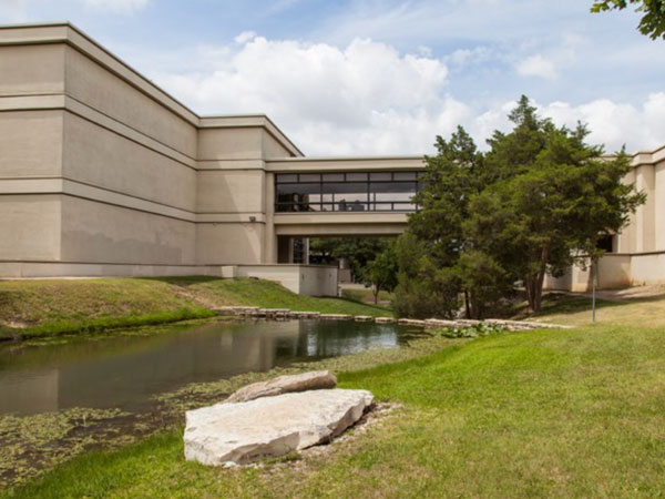 Photo of the W building and the campus creek that runs though the middle of campus. You can see a limestone chalk rock and cypress trees along the shore of the creek.