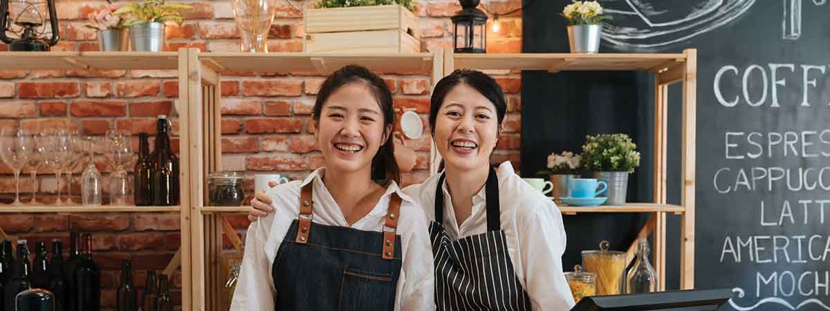 Two successful young baristas women standing in bar counter in cafe in aprons smiling confidently to camera in coffee shop.