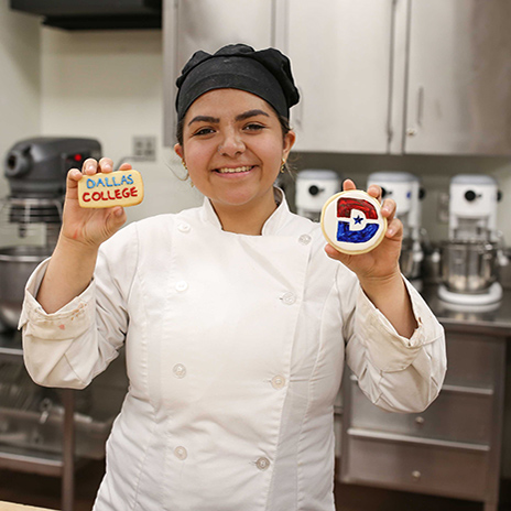 Culinary student smiling holding two delicious sugar cookies decorated with Dallas College logos and colors