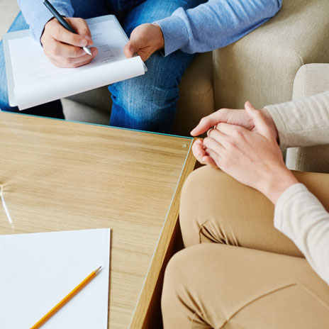 Two people sit around a table, only their laps are seen as one person writes on a notepad and the other sits with their hands folded in their lap.
