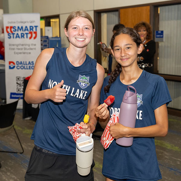 Two individuals wearing North Lake athletic shirts holding snacks and drinks at a Dallas College event, with a Smart Start banner visible in the background.