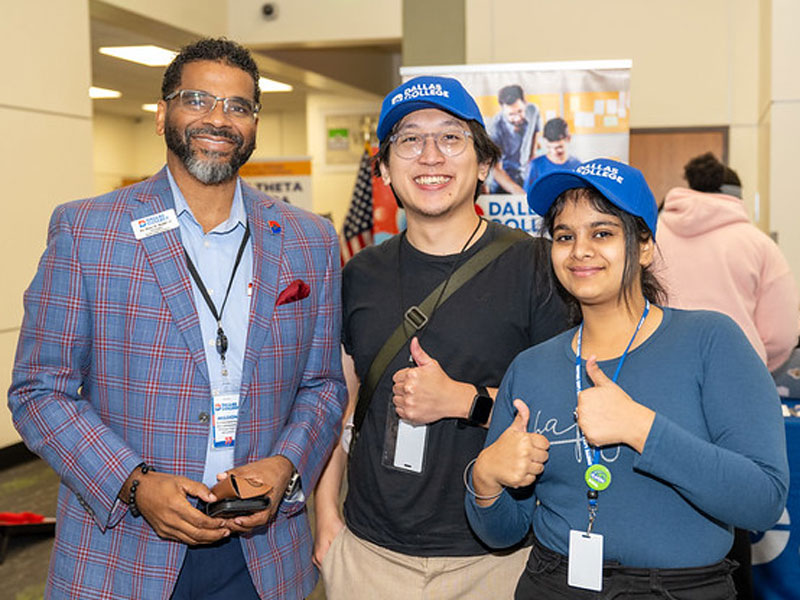 Three people at an indoor event wearing badges and blue Dallas College caps, with a banner in the background.