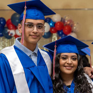 Two Dallas College graduates in blue caps and gowns standing together with balloons in the background.