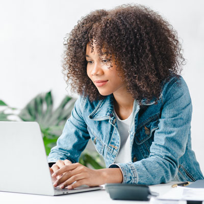 A woman with curly hair in a denim jacket focused on a laptop at a white desk, surrounded by plants, conveying a calm and productive atmosphere