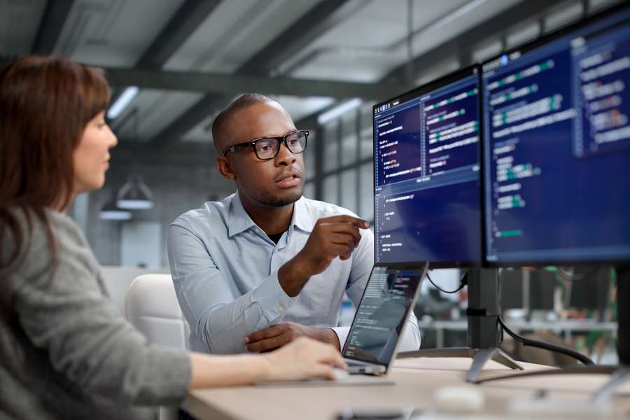 male and female coworker sitting in front of a computer monitor with a laptop