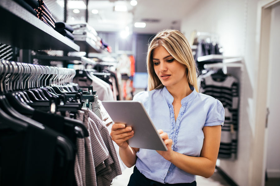 business woman in light blue top looking at clipboard with clothes hanging on a rack in the background