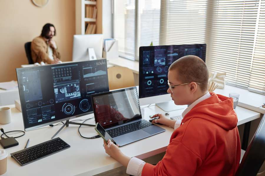 worker in a red hoodie sitting at a computer desk with computer monitors and a laptop