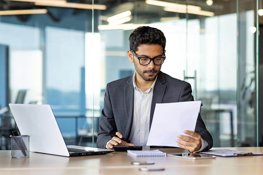 Business professional sitting at a desk in a modern office, reviewing documents while working with a laptop, calculator and notebook nearby.