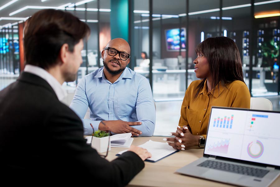 Three business professionals sitting around a conference table in a modern office, discussing documents with a laptop displaying charts nearby.