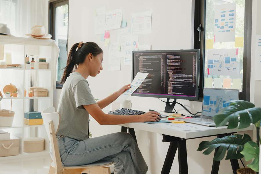 female sitting at a desk holding a paper in front of computer monitors