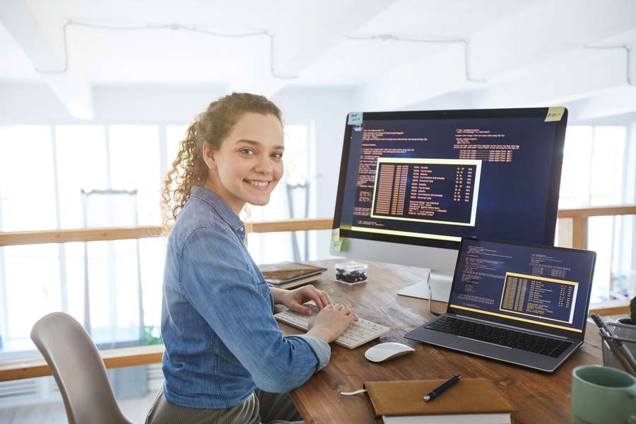 female sitting at a computer desk with laptop and looking at camera smiling