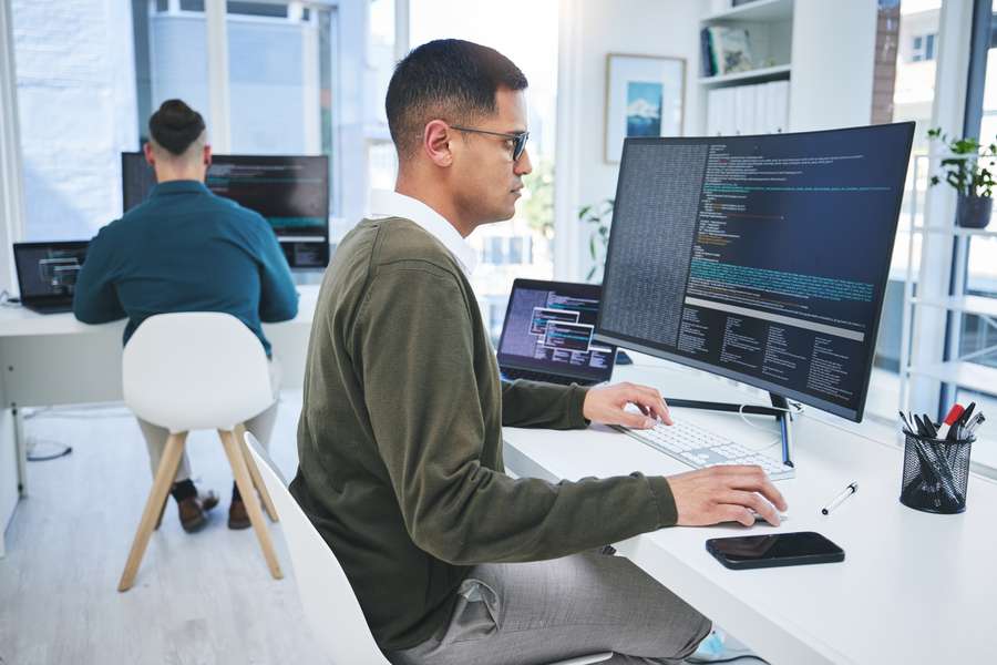 male worker in a green sweater at a computer desk looking at a monitor with code