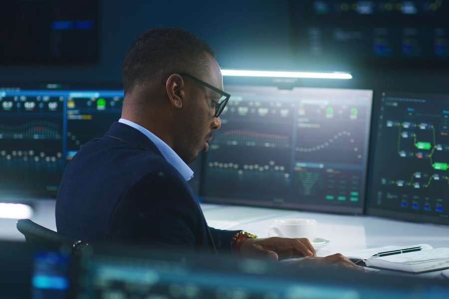 male in a blue suit sitting at a computer desk looking down at a notebook