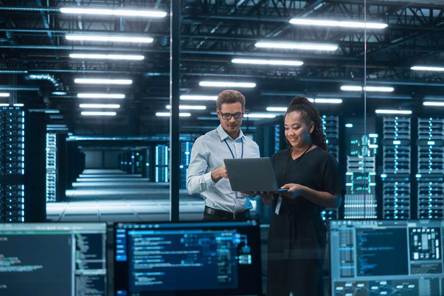 male and female coworkers standing in a room filled with computers and holding a laptop