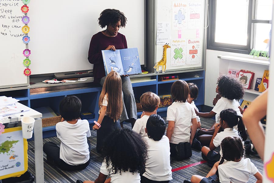 female teaching standing in front of the classroom holding a storybook with a group of children