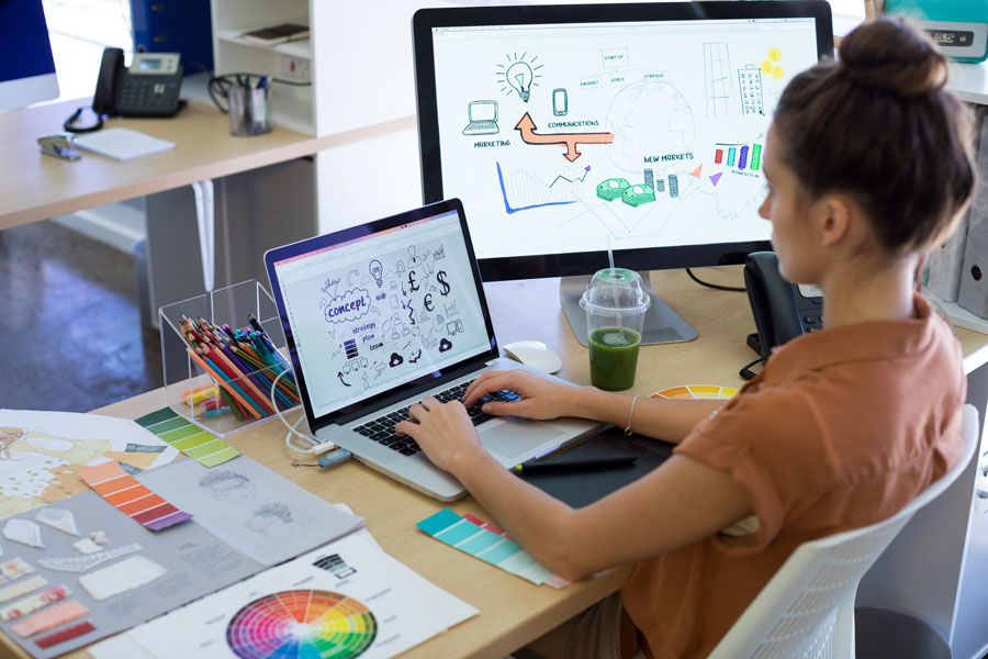 female sitting at desk typing on laptop with large monitor