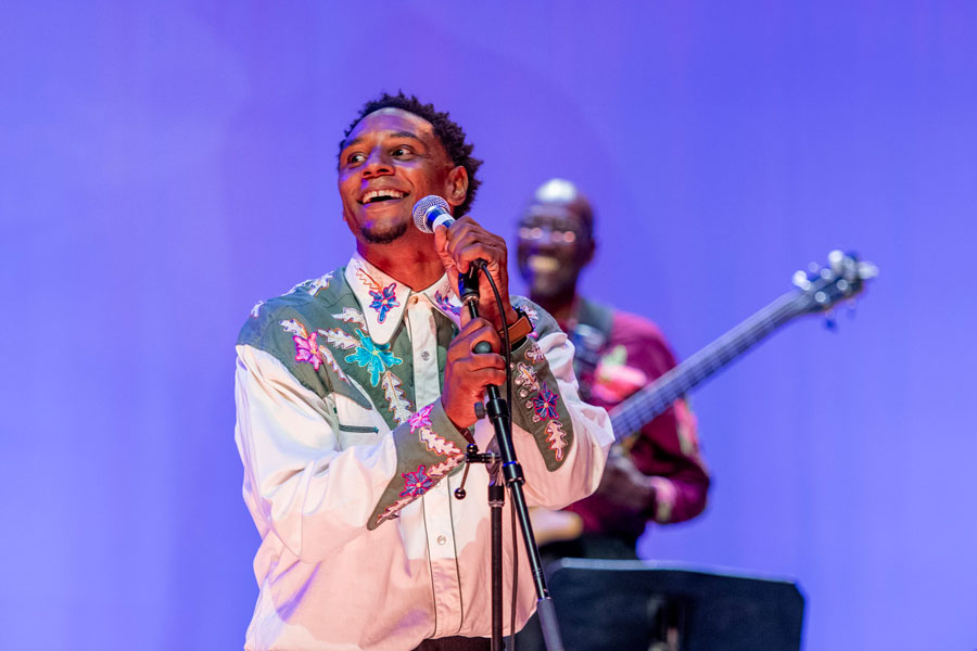 male performer on stage holding a microphone and singing in front of a purple background