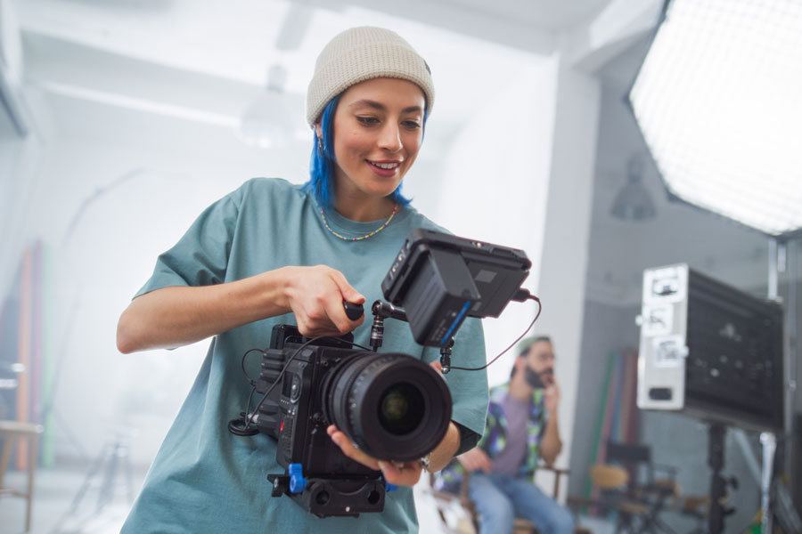 Young Female Cinematographer With Blue Hair Adjusts Professional Camera In Studio, Showcasing Teamwork In Creative Film Production Environment, With Colleagues Collaborating In Background.