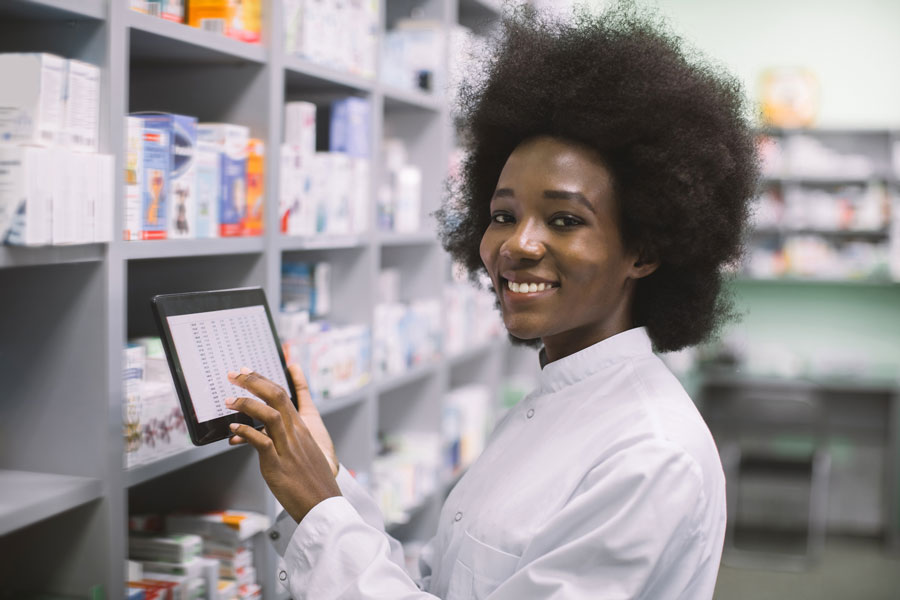 Young female pharmacist smiling and using digital tablet in front of pharmacy shelves.