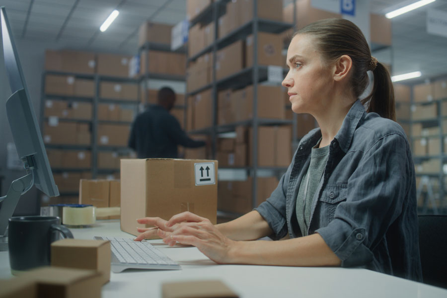 Female warehouse clerk using computer and typing on keyboard with brown box nearby