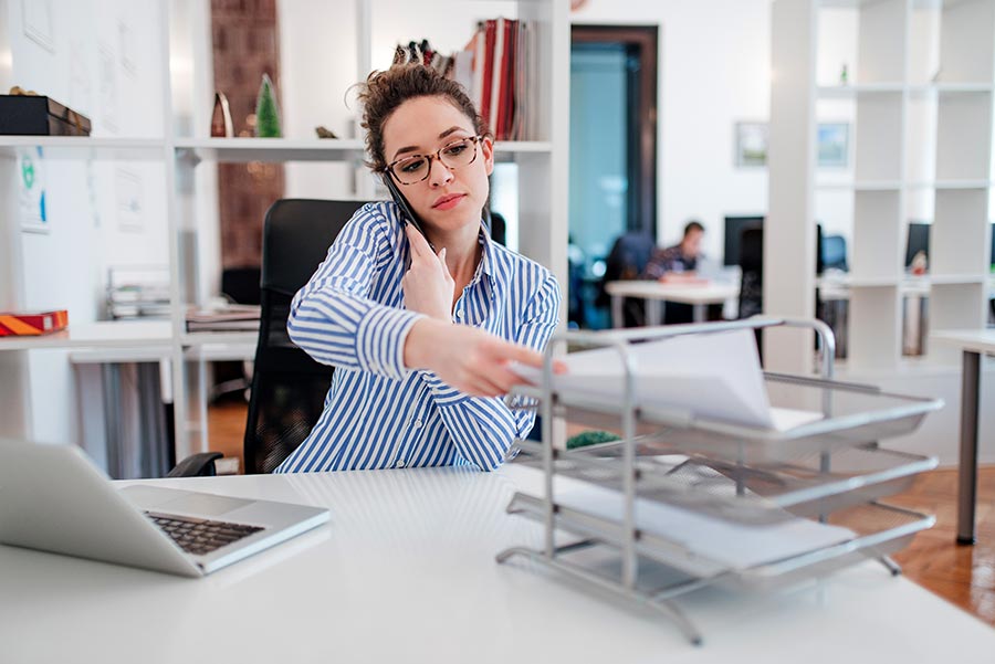 Office employee talking on the phone while reaching for paperwork on a document tray, with a laptop and open office workspace in the background.