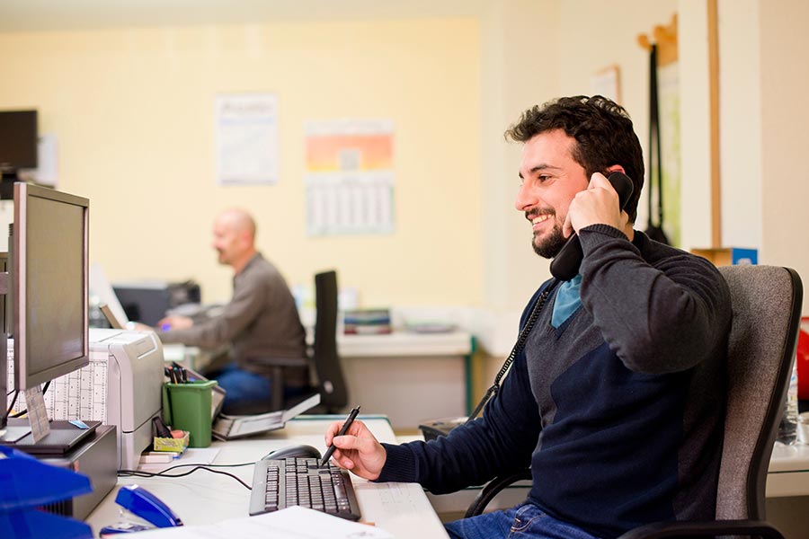 Man working at a desk in an office, smiling while talking on the phone, writing notes and using a computer with another employee in the background.