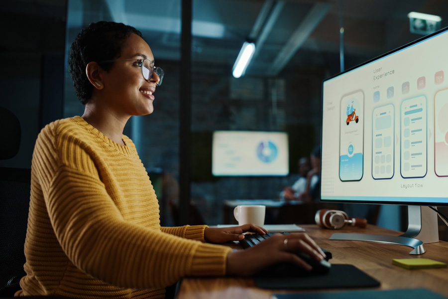 Person working at a desk on a large monitor displaying mobile app user interface designs, with a keyboard, mouse, coffee mug, and headphones on the desk in a modern office setting.