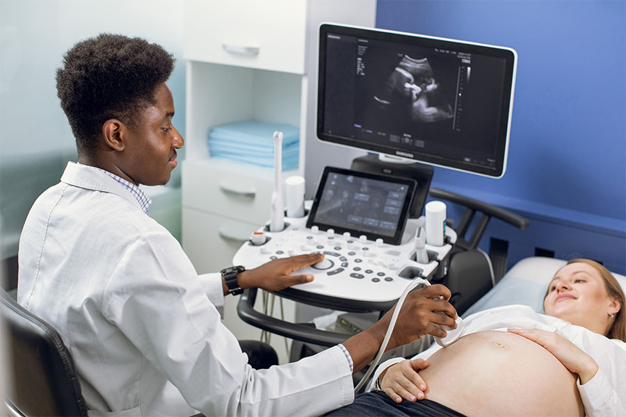 Medical professional performs an ultrasound on a pregnant patient; fetus image is visible on the monitor, with faces blurred for privacy.