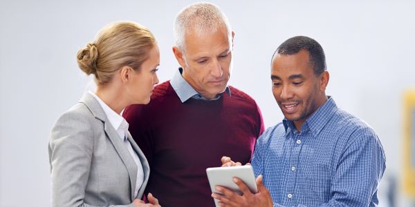 Three employees stand in a warehouse and review info on a tablet