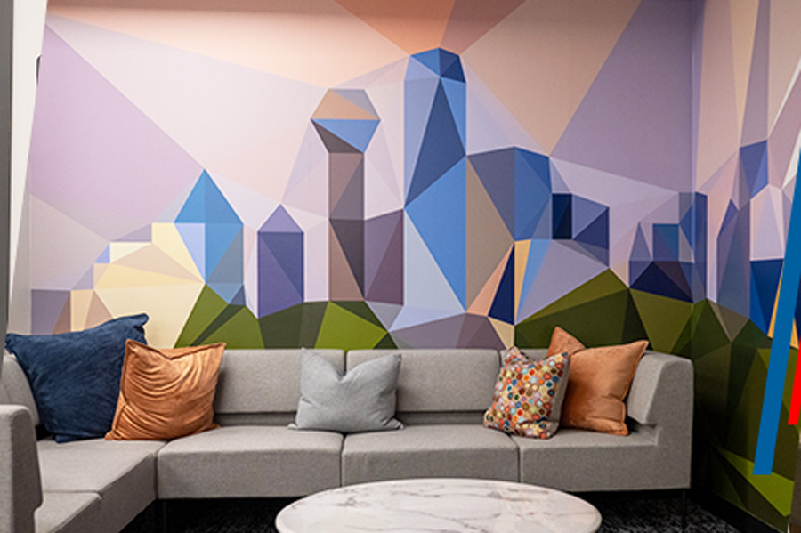 Modern lounge area with a grey sectional sofa decorated with pillows in blue, orange, light grey, and multicolored floral patterns. Behind the sofa is an abstract mural featuring geometric shapes in blue, purple, green, and yellow. A round marble coffee table sits in front of the sofa, creating a contemporary and vibrant setting.