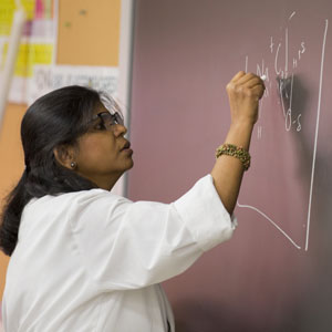A woman in a white lab coat writes scientific equations on a chalkboard