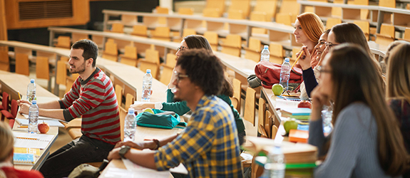 Students sit in desks in a large, mostly empty classroom