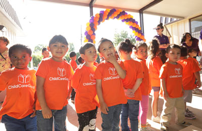 Group of children in matching orange 'ChildCareGroup' shirts under a purple and yellow balloon arch.