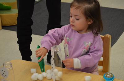 Child in a purple sweatshirt using green tongs to move cotton balls between plastic bottles with paper faces attached.