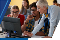 a smiling older woman leans over two young people sitting at a computer workstation