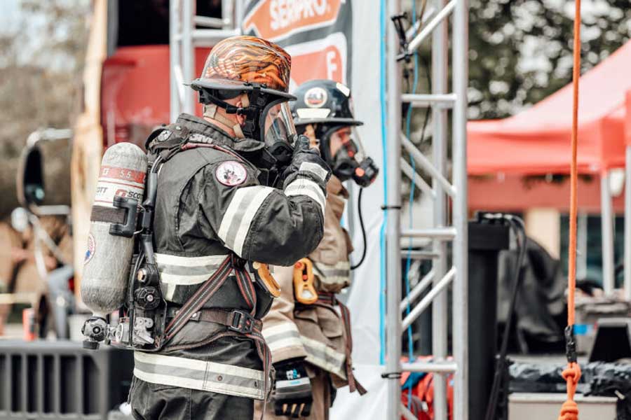 Two firefighters outdoors in full protective gear, including helmets, face masks, and oxygen tanks. The firefighter in the foreground is adjusting their face mask with a gloved hand. Behind them, a metal structure and a red tent are visible.