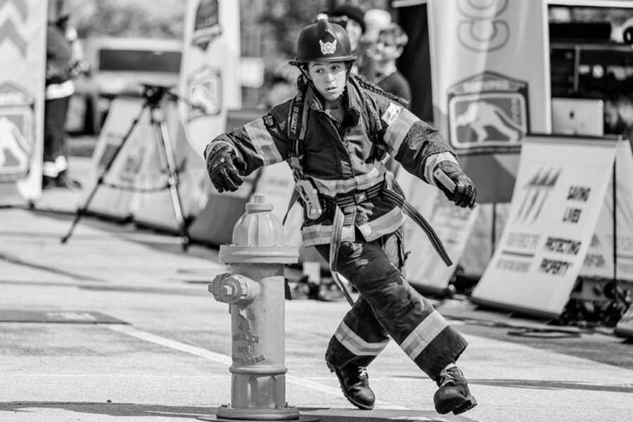Black and white photo of a firefighter in full protective gear navigating around a fire hydrant during a competition or training exercise. The firefighter has one leg lifted and arms extended for balance. In the background, banners, firefighting equipment, and spectators are visible.