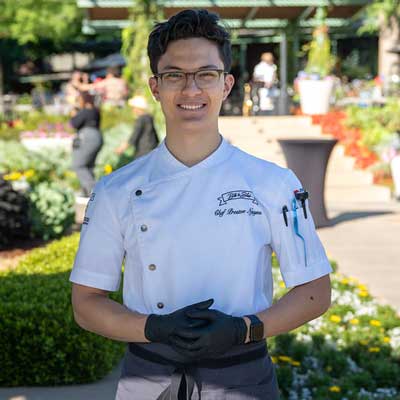 Chef Preston Nguyen standing outdoors in a garden setting, wearing a white chef's jacket.