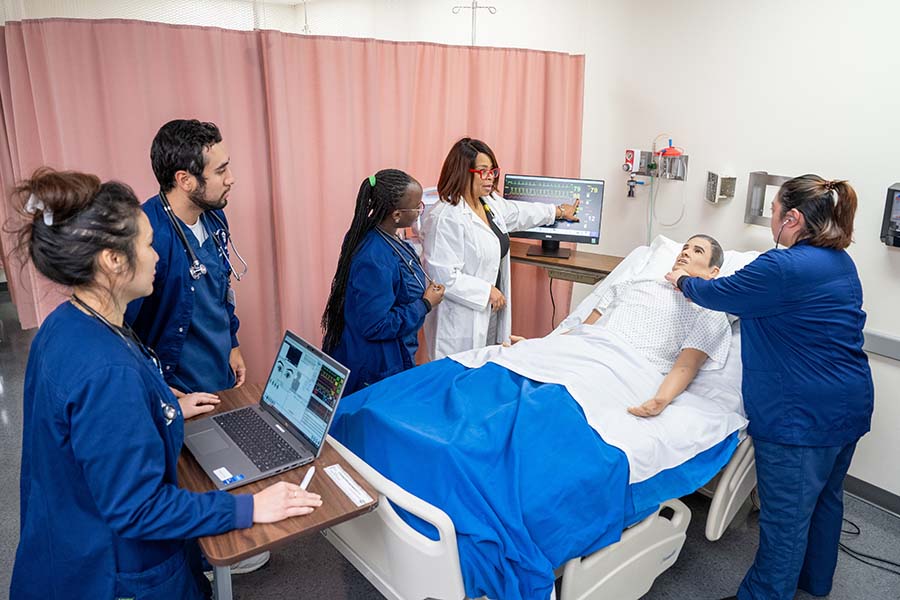 Group of five nursing students with instructor gathered around manikin in bed.
