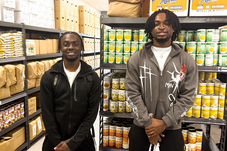 Two students stand in front of campus food pantry shelves.