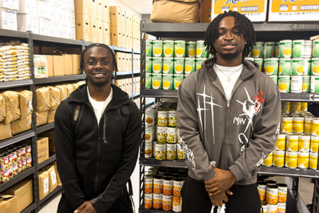 Two students stand in front of campus food pantry shelves.