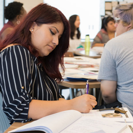 Group of students collaborating at a table, with one person writing in a notebook during a study session or classroom activity