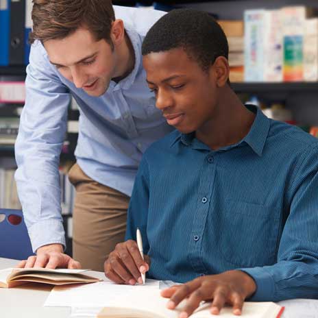 Student writing in a book while receiving guidance from another person in a study environment