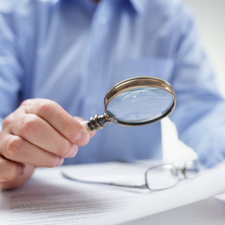 Person holding a magnifying glass over a document with eyeglasses nearby