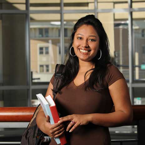Student holding books and shoulder bag indoors, standing near large windows