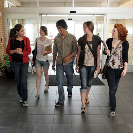 Group of students walking and talking in a school hallway near an entrance, carrying books and bags.