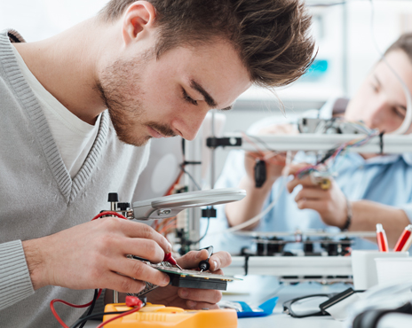 Two individuals working on electronic circuit boards at a workbench, with tools and components visible.