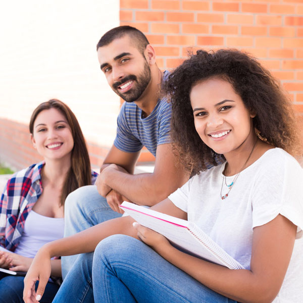 Three students sitting outdoors on campus, holding notebooks and engaged in academic discussion