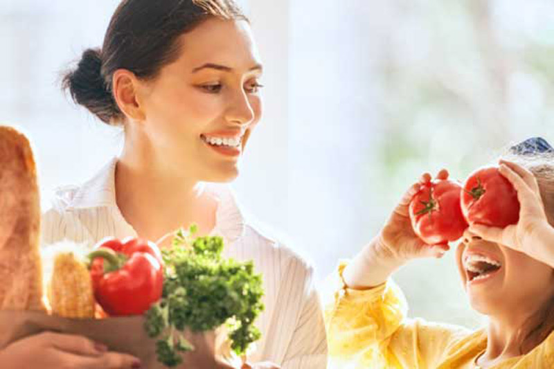 Mother and daughter with groceries, one playfully holding tomatoes over their eyes.