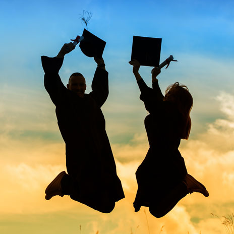 Two graduates in caps and gowns jumping joyfully against a vibrant sunset sky, celebrating their achievement